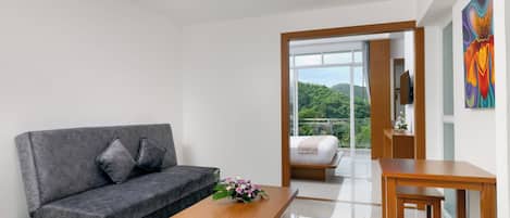 A modern hotel room with a grey sofa, a wooden coffee table, a bed with a view, and a painting on the wall.