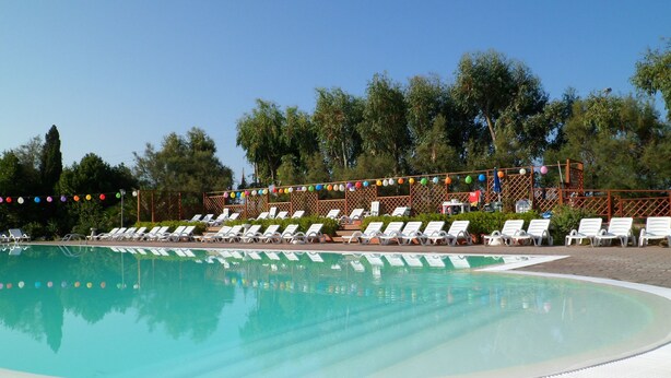 A swimming pool with white lounge chairs arranged around it, a wooden deck, and a backdrop of trees. - New Camping Le Tamerici (Cecina)