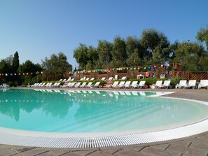 Una piscina con sillones blancos dispuestos a su alrededor, una terraza de madera y árboles como fondo.