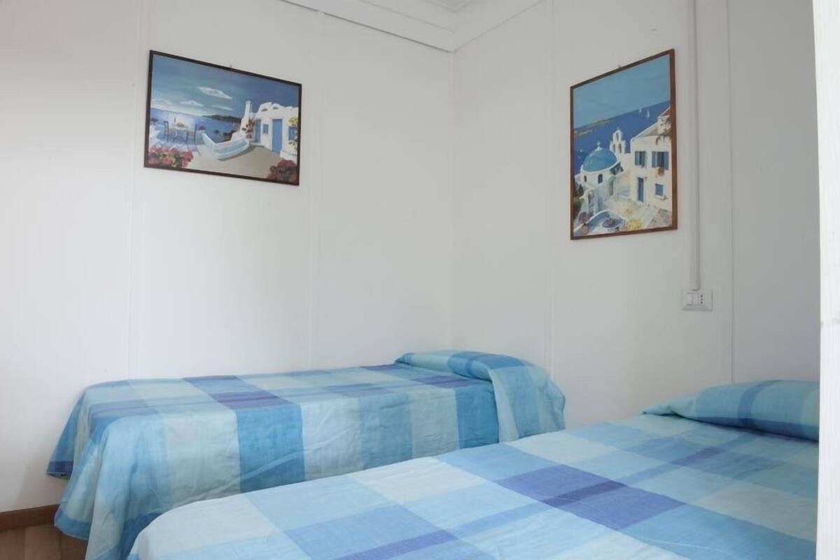 A room with two beds, each with a blue checkered bedspread, and two paintings of coastal scenes on the walls.