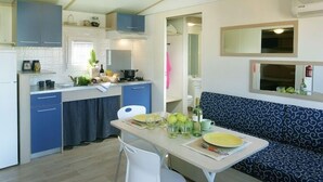 A compact kitchen with blue cabinets, a dining table set with plates and glasses, and a blue patterned sofa in the living area. - New Camping Le Tamerici (Cecina)