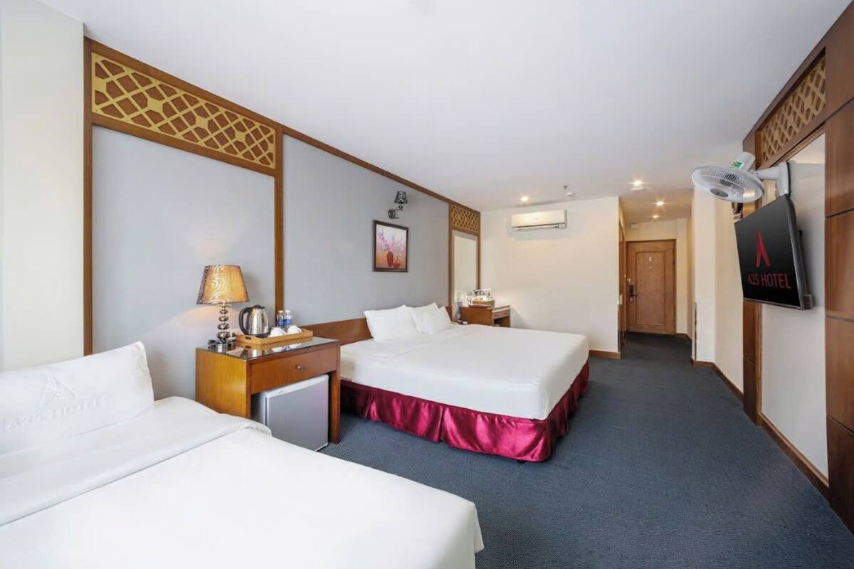 A hotel room with two beds, a desk with a lamp, a small refrigerator, and a TV on the wall.
