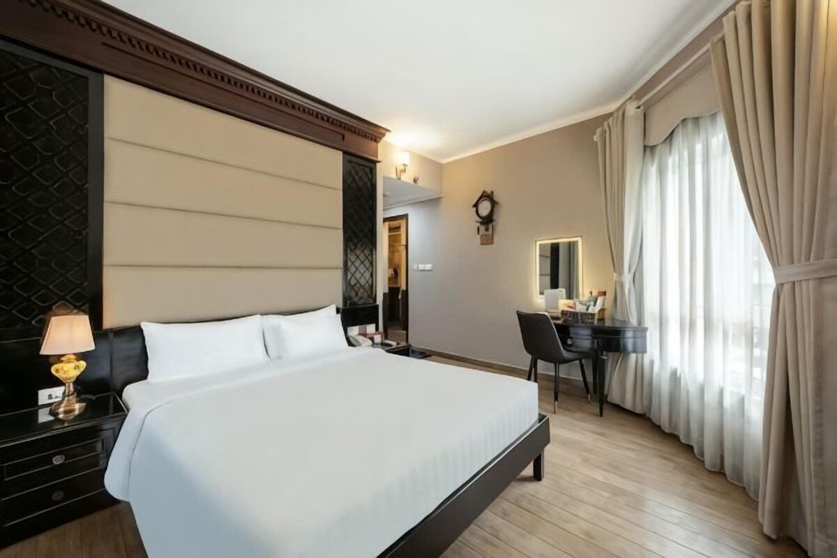 A hotel room with a large bed, a desk, a chair, a lamp, and a window with curtains.
