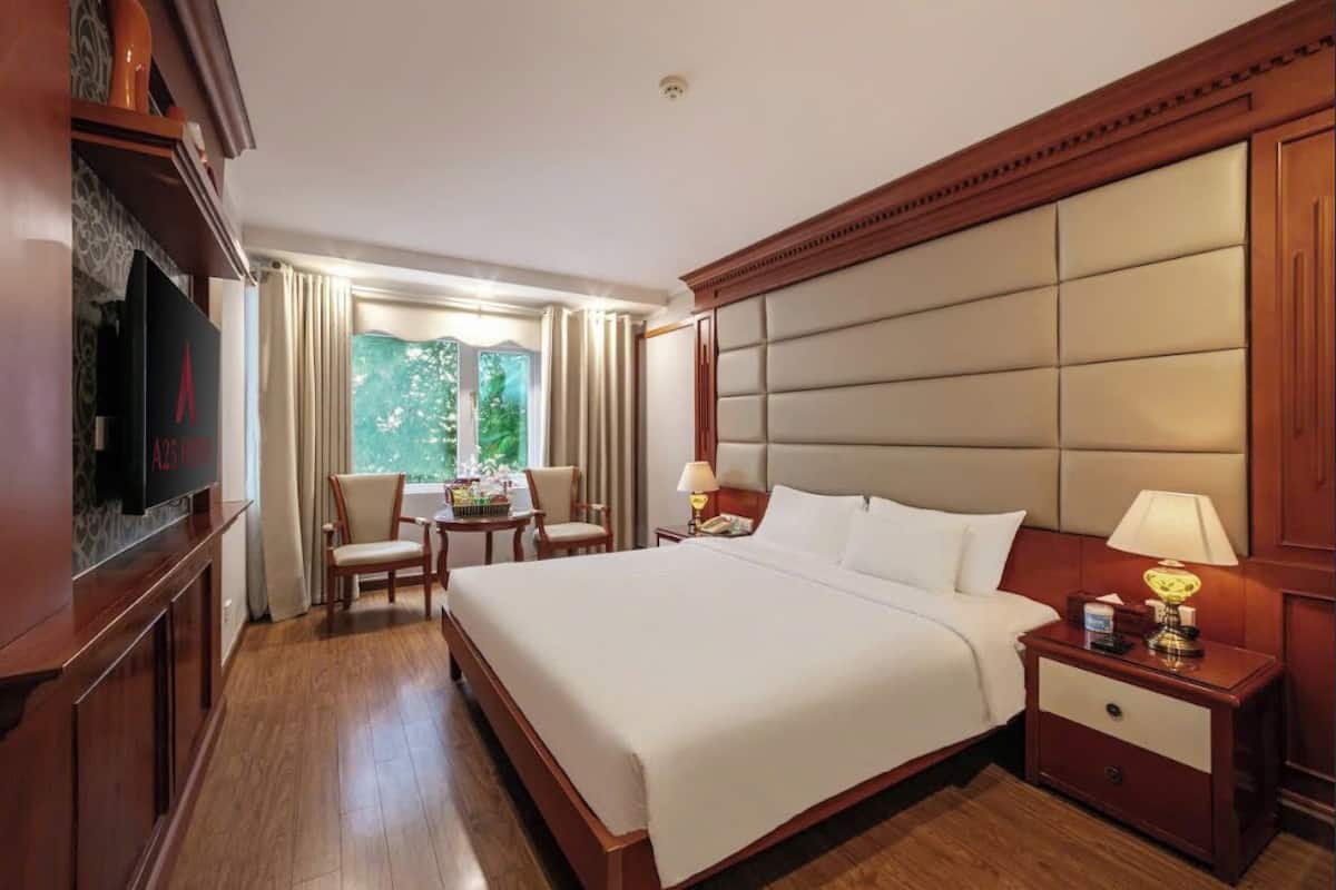 A hotel room with a large bed, wooden headboard, a television on a cabinet, a small table with chairs, and a window with curtains.