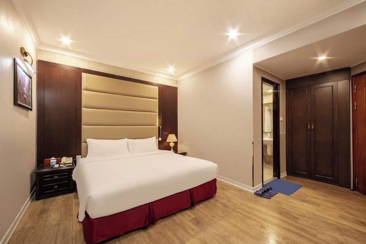 A hotel room with a large bed, wooden headboard, bedside tables with lamps, a wooden floor, and a door leading to a bathroom.