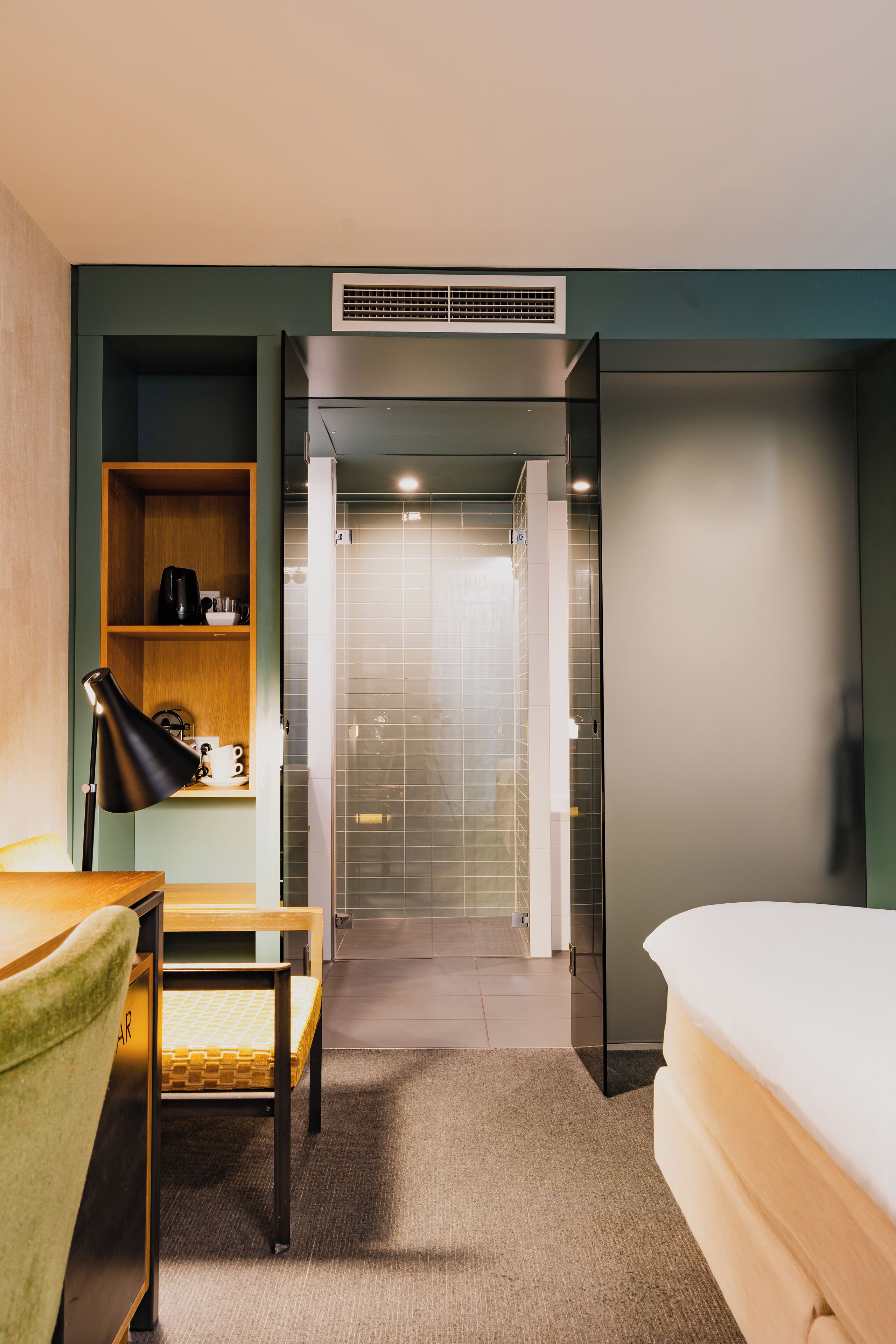 A hotel room with a bed, a desk with a lamp, a shelf with toiletries, and a shower area.