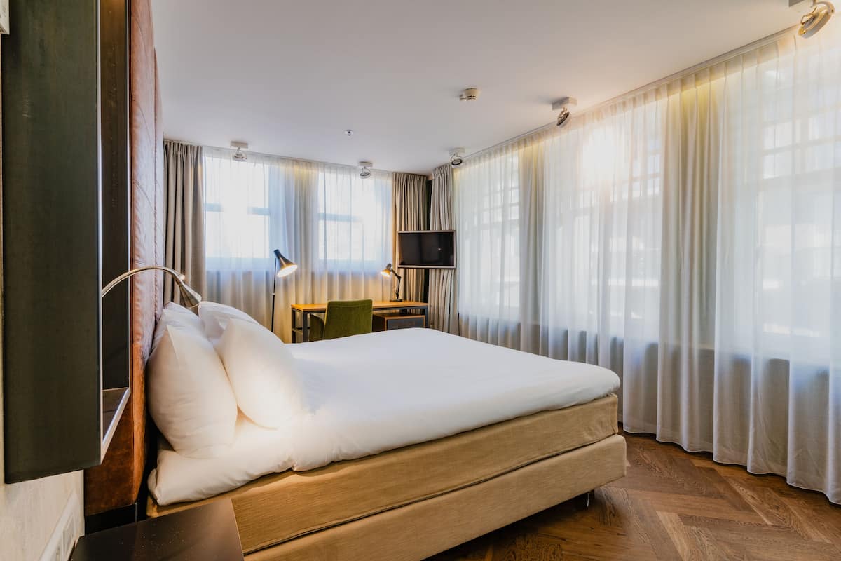 A hotel room with a large bed, a desk with a chair, a television, and sheer curtains.