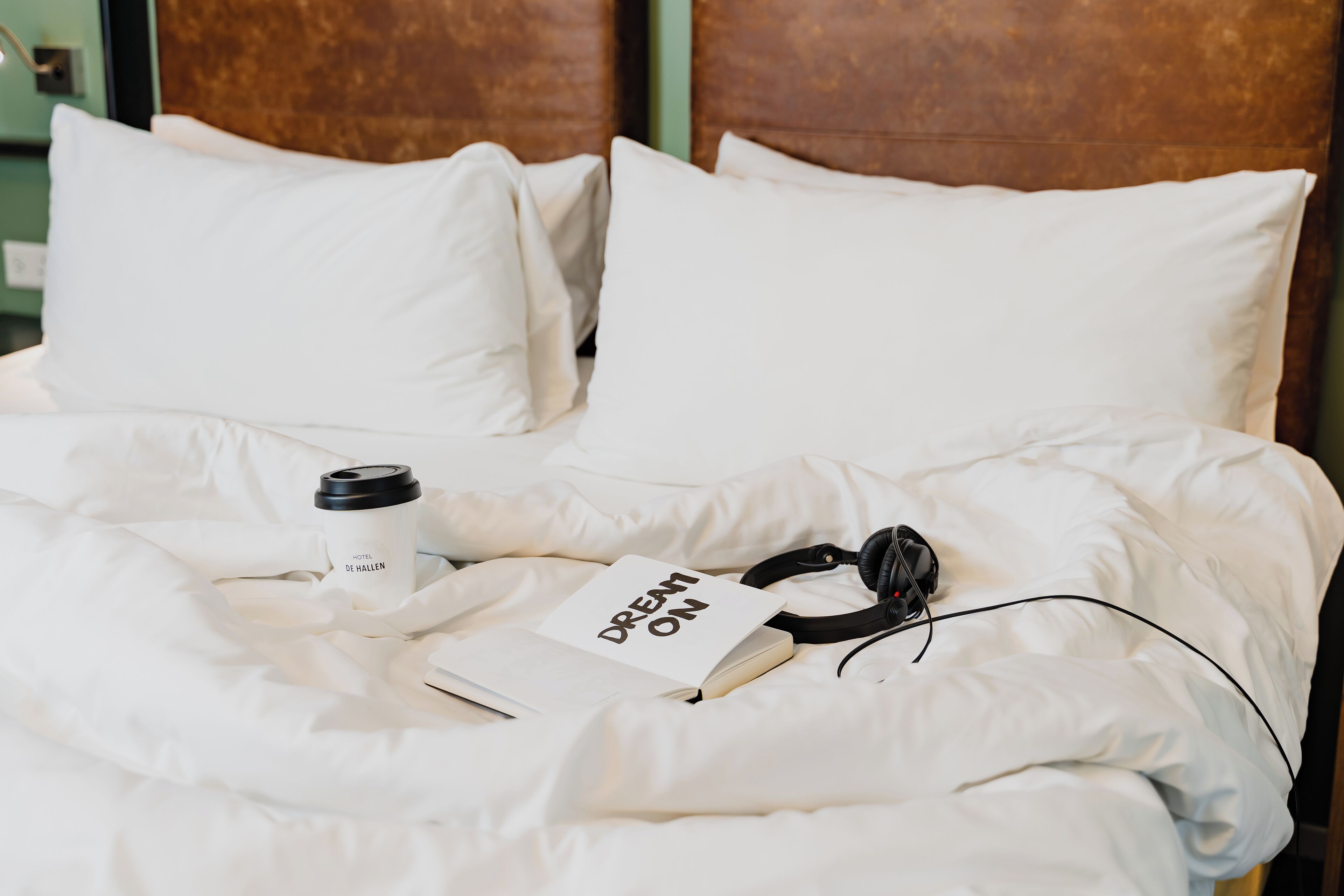 Una cama bien hecha con ropa de cama blanca, un libro titulado 'DREAM ON', unos auriculares negros y una taza de café con la etiqueta 'HOTEL DE HALLEN'.