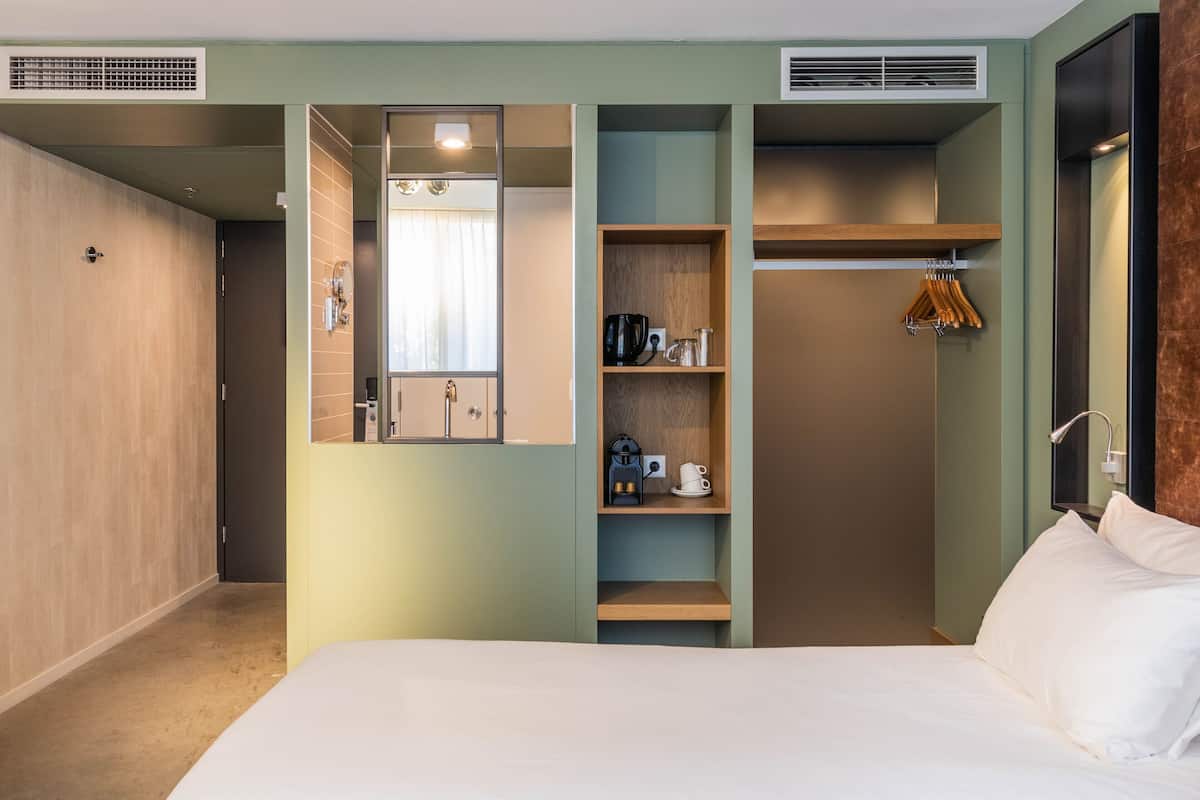 A hotel room with a bed, a wardrobe, a mirror, a shelf with toiletries, and a hanging rack.