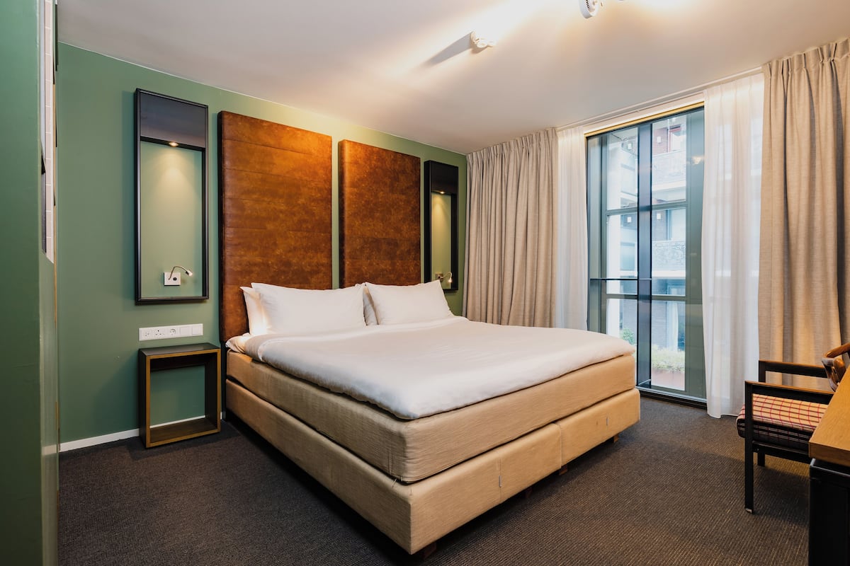 A hotel room with a large bed, a small table, a chair, a window with curtains, and a wall-mounted mirror.