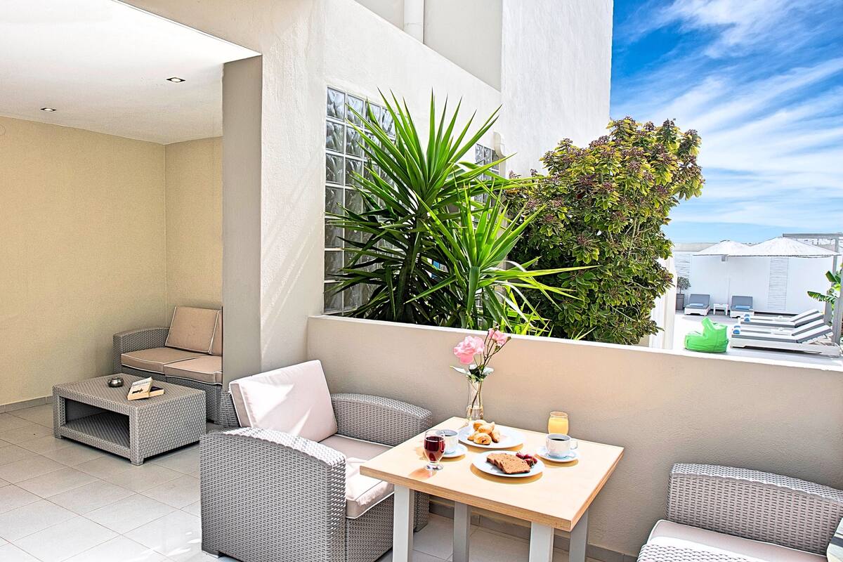 Een modern balkon met rieten meubilair, een eettafel met ontbijt en uitzicht op de stad.