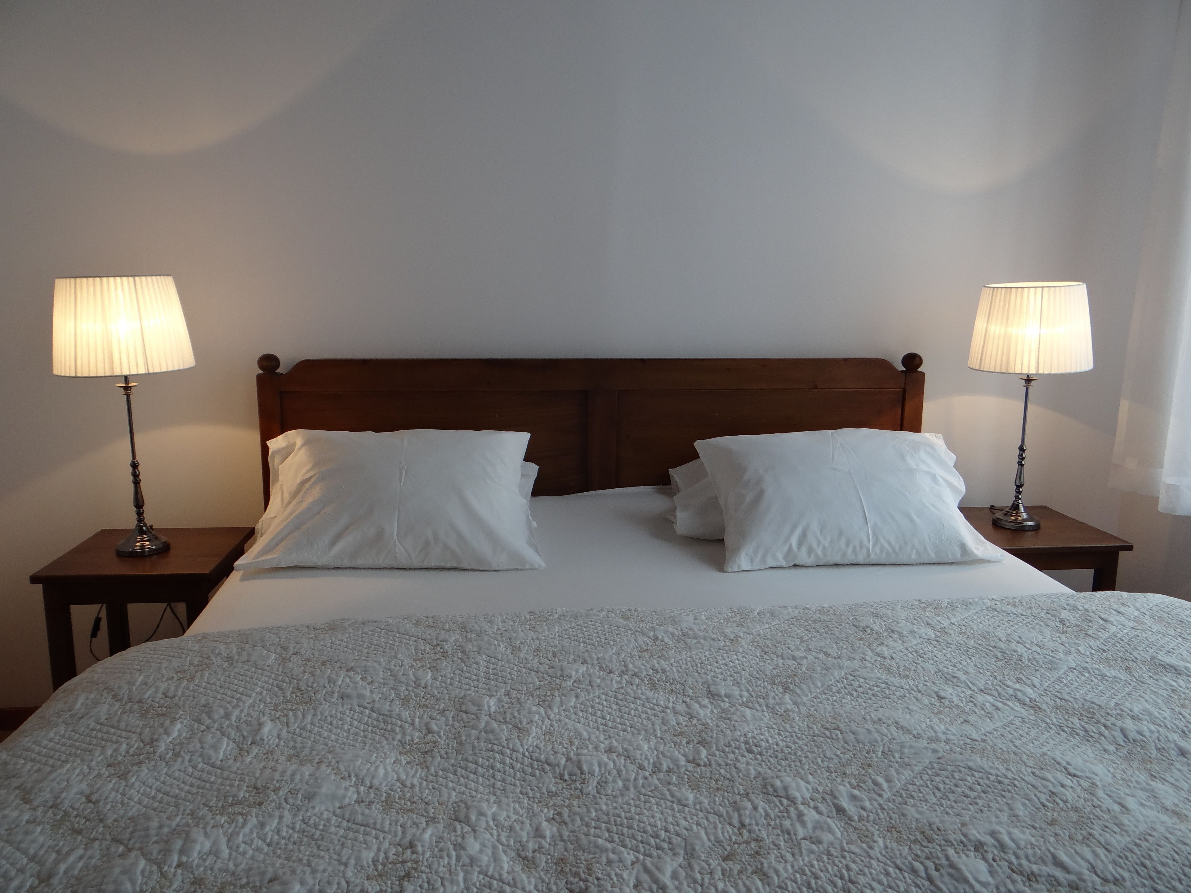 A neatly made bed with white linens and pillows, flanked by two bedside tables with lamps, in a room with a wooden headboard and plain walls.