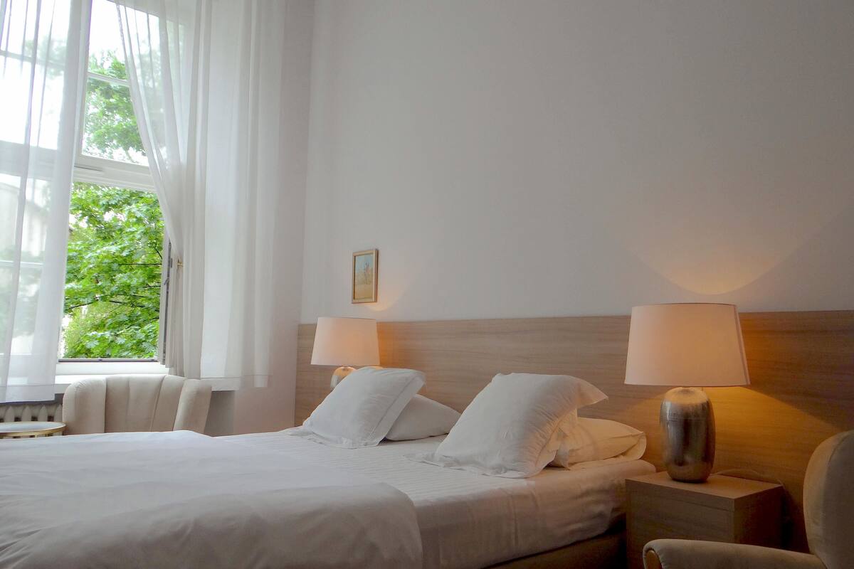 Une chambre d’hôtel avec un grand lit, deux lampes de chevet, une petite table et une vue sur la verdure par la fenêtre.