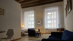 En stue med sofa, lænestol, lille bord og lampe. To vinduer med gardiner, en radiator og en lysekrone.