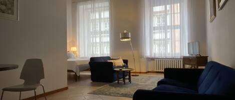 A living room with a sofa, a chair, a small table, and a lamp. There are two windows with curtains, a radiator, and a chandelier.