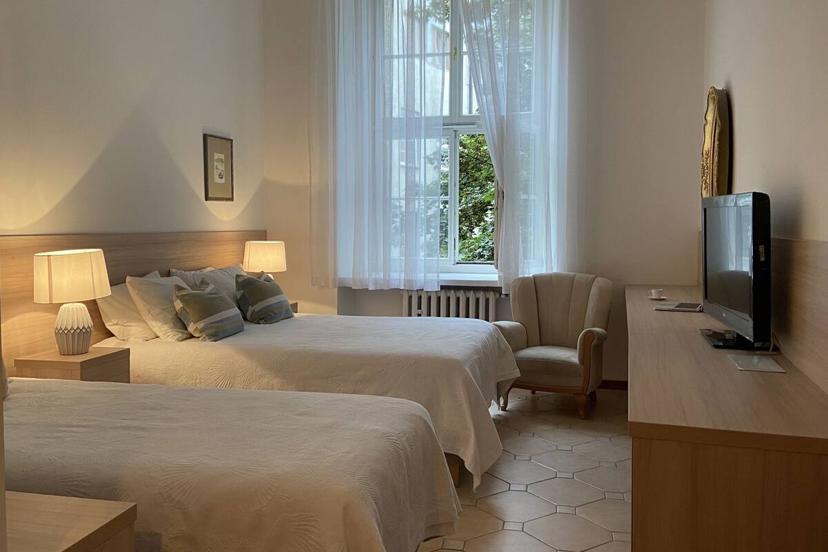 Une chambre d’hôtel avec deux lits, une chaise, un téléviseur, une fenêtre avec des rideaux légers et un radiateur.