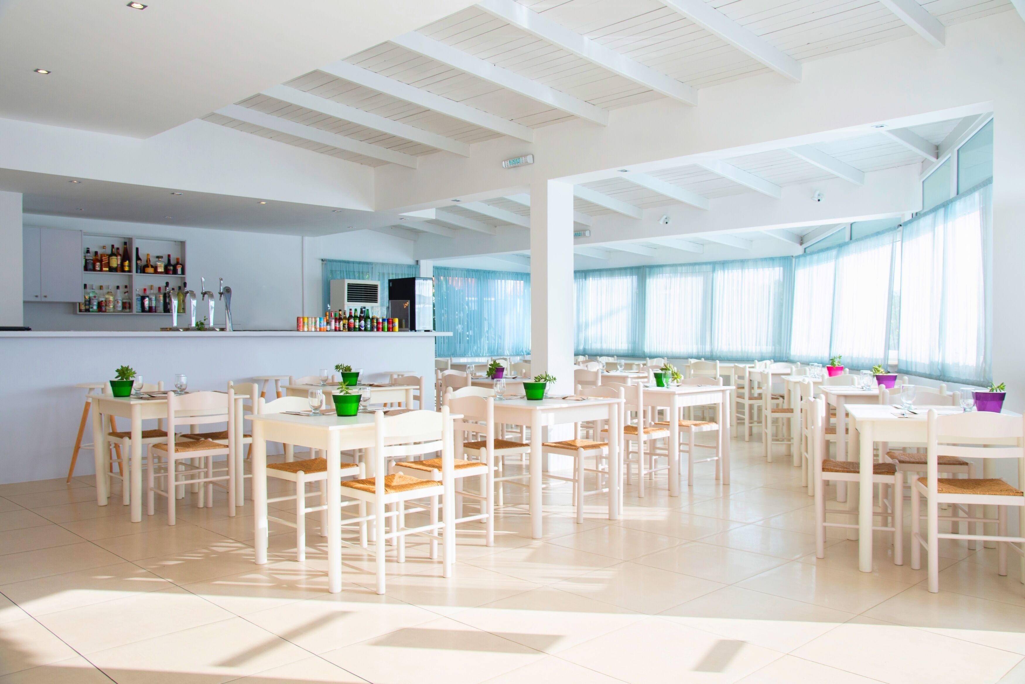 A spacious dining area with white tables and chairs, a bar with shelves of bottles, and large windows with blue curtains.