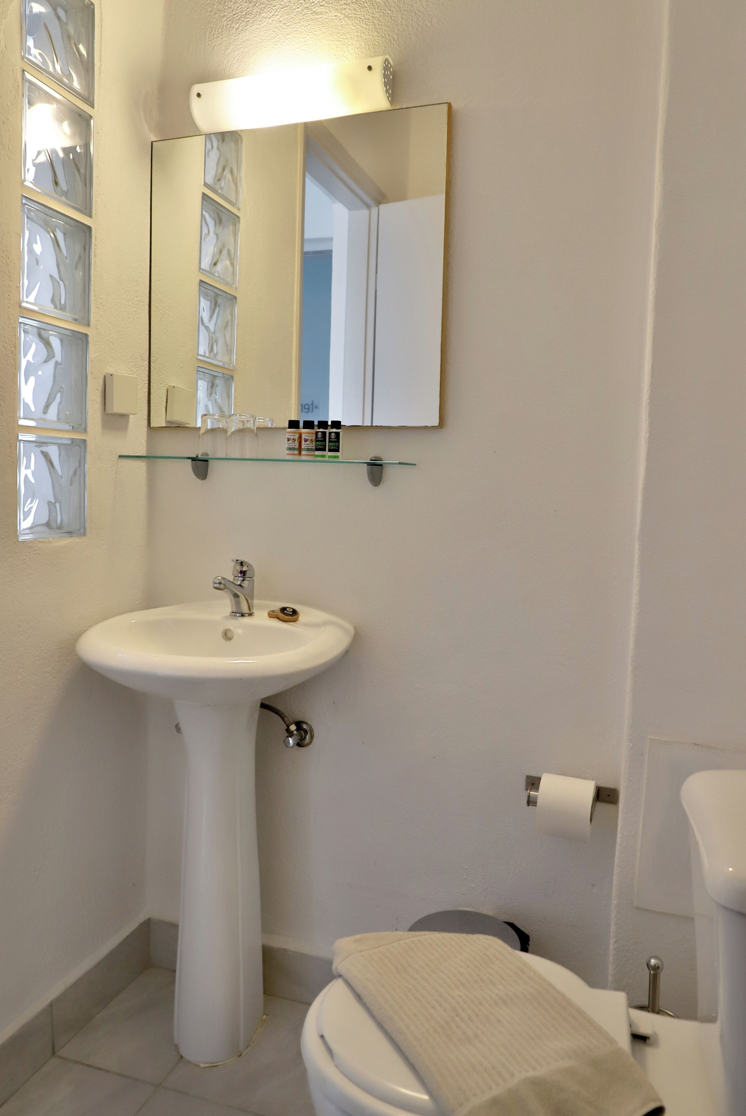 A bathroom with a pedestal sink, a mirror, a glass shelf with toiletries, and a toilet.
