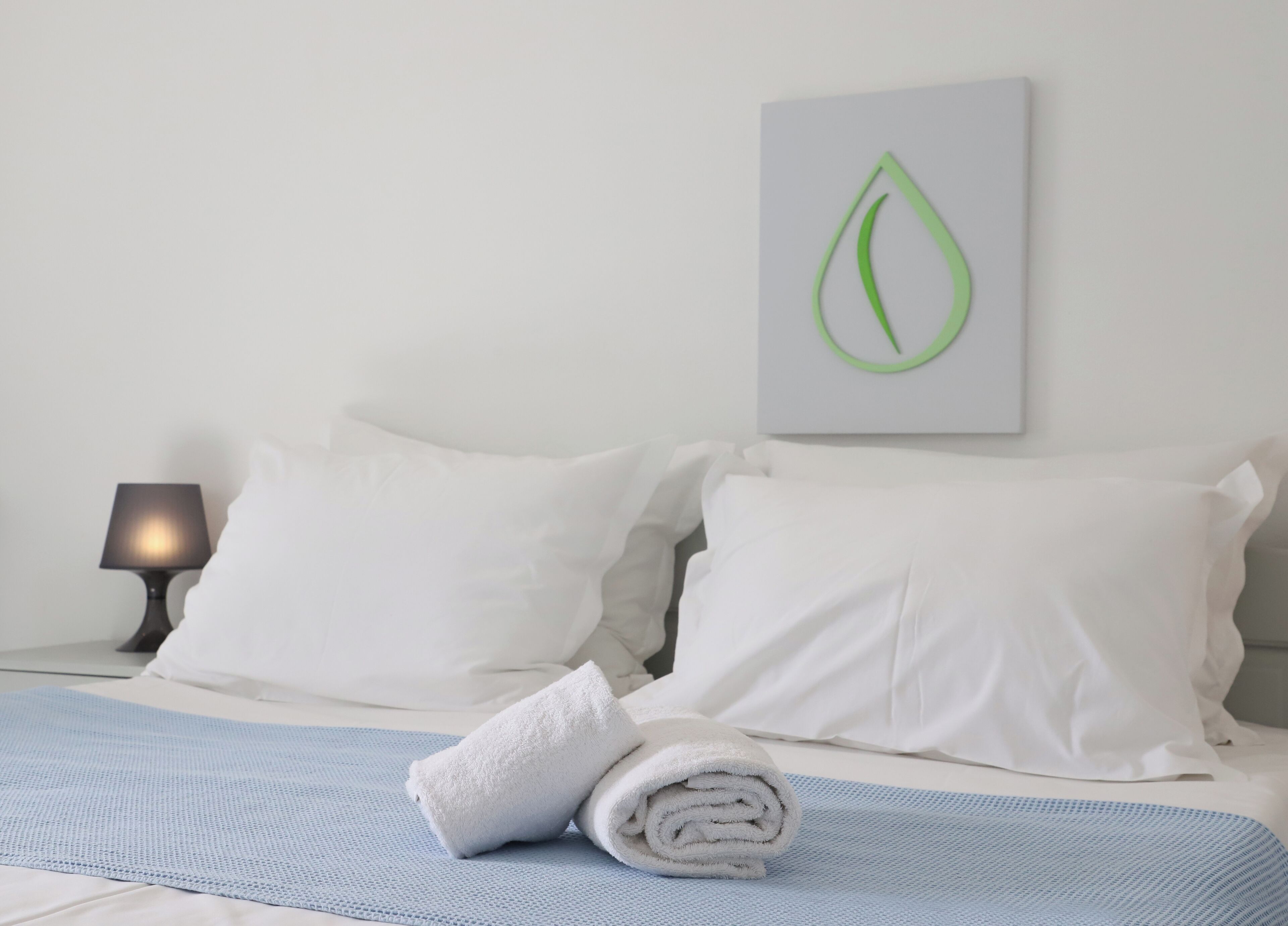 Una cama bien hecha con ropa blanca, una toalla blanca enrollada, un tapete azul con textura y una obra de arte con forma de hoja verde montada en la pared.