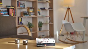 Una habitación con una mesa de madera, dos candelabros metálicos, una pila de libros, una mecedora blanca y una lámpara.