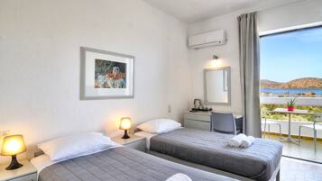 Une chambre d’hôtel avec deux lits, un bureau, une chaise, une petite table avec un pot de plante et un balcon avec vue sur la mer.