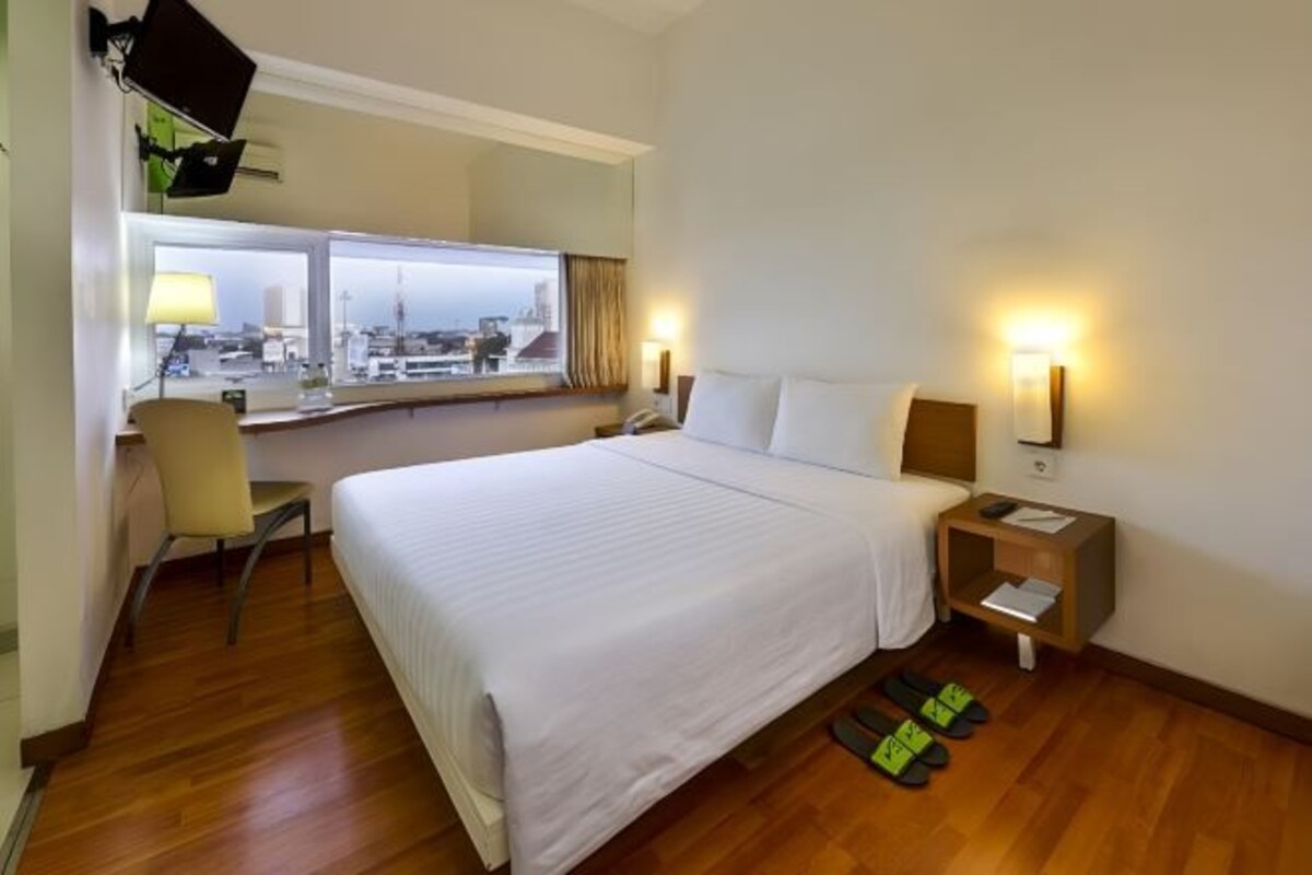 A hotel room with a large bed, a desk with a chair, a window with a view, and a nightstand with a lamp.