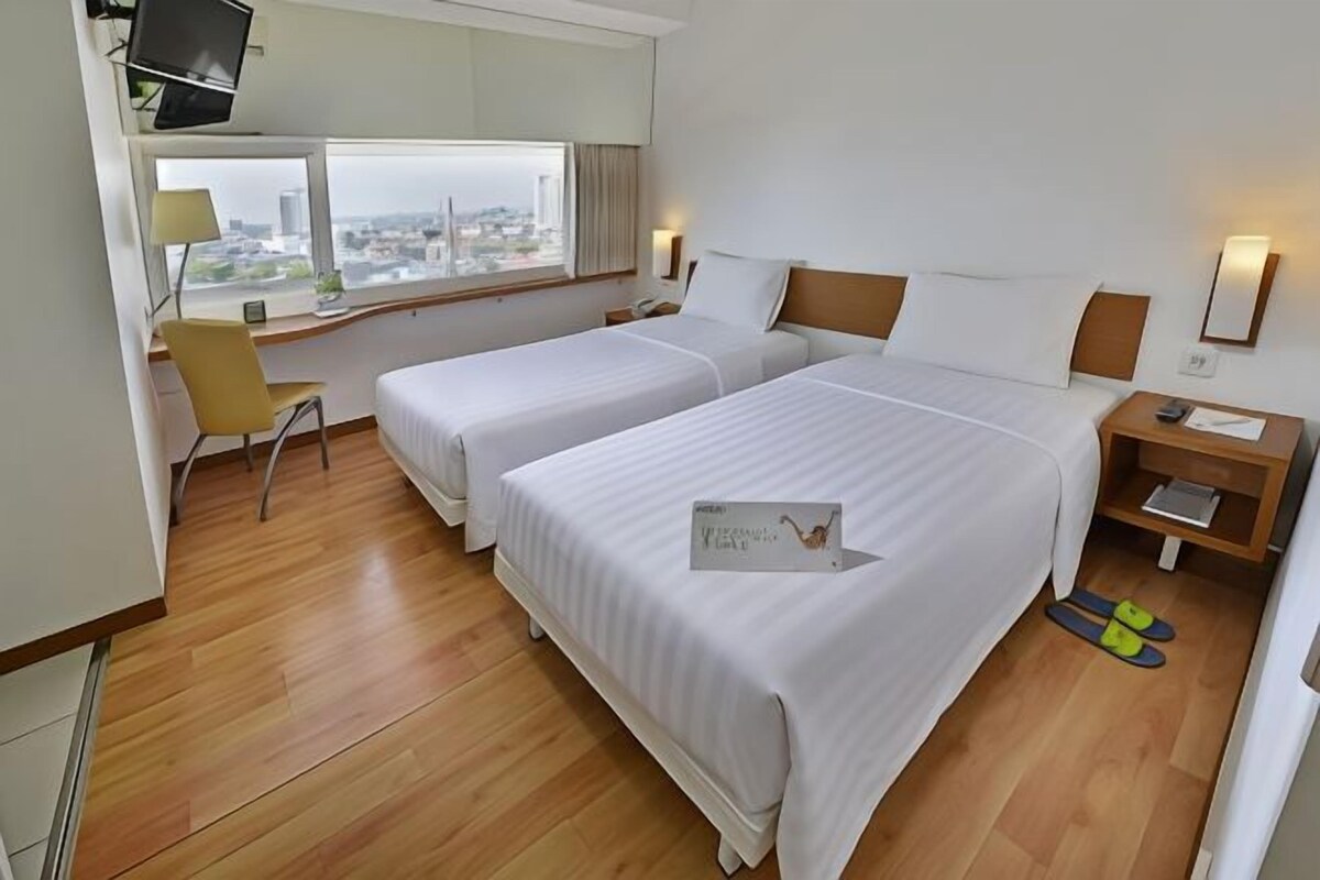 A hotel room with two beds, a desk, a chair, a TV, and a window with a city view.