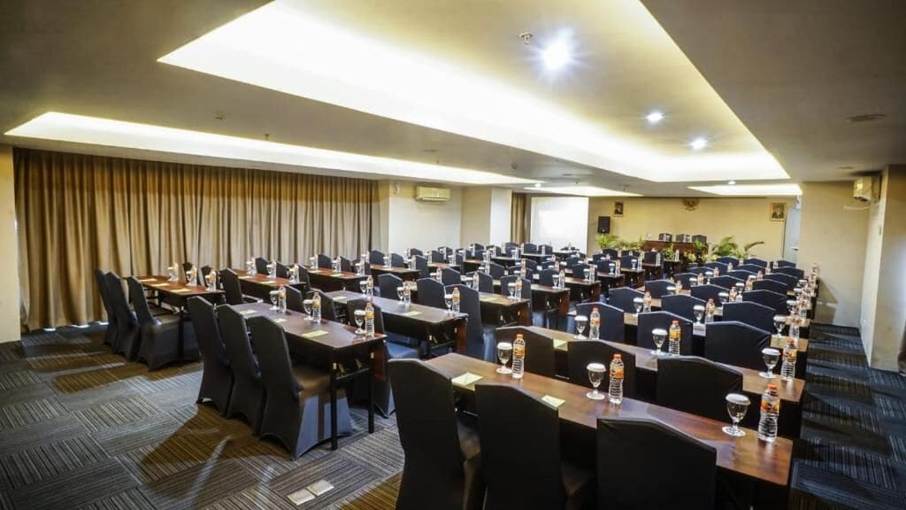 A conference room with rows of tables and chairs, equipped with water bottles and glasses, and a large screen at the front.