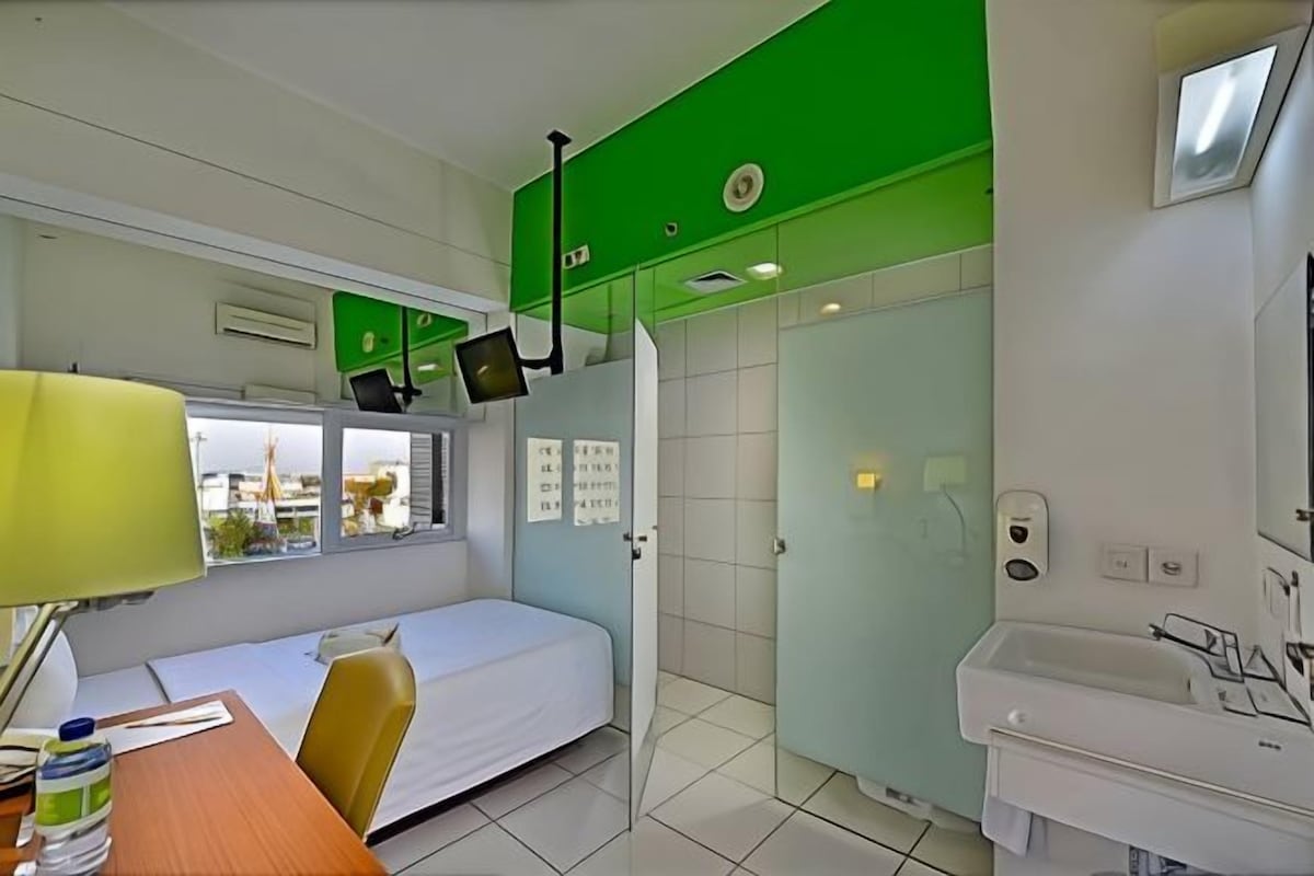A hotel room with a bed, a desk with a lamp, a bathroom with a sink, and a green accent wall.