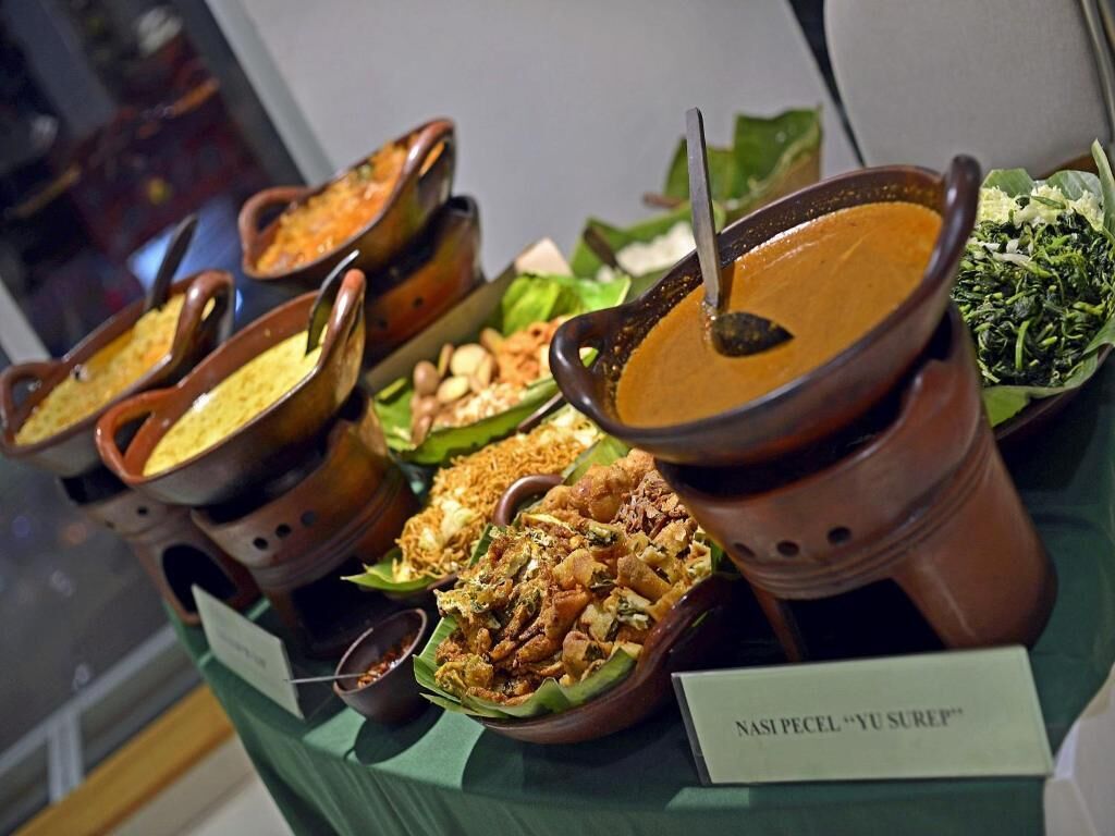 A variety of dishes displayed in traditional clay pots on a table, with a sign indicating 'Nasi Pecel' and 'Yu Sreep'.
