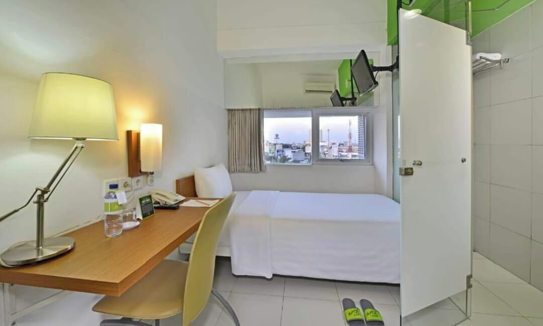 Una habitación de hotel con cama, escritorio con lámpara, silla, ventana con vista y un par de pantuflas en el suelo.