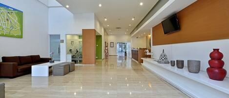 A spacious lobby with a modern design, featuring a large seating area with a brown sofa and a white coffee table, a long reception desk, and a wall-mounted TV.