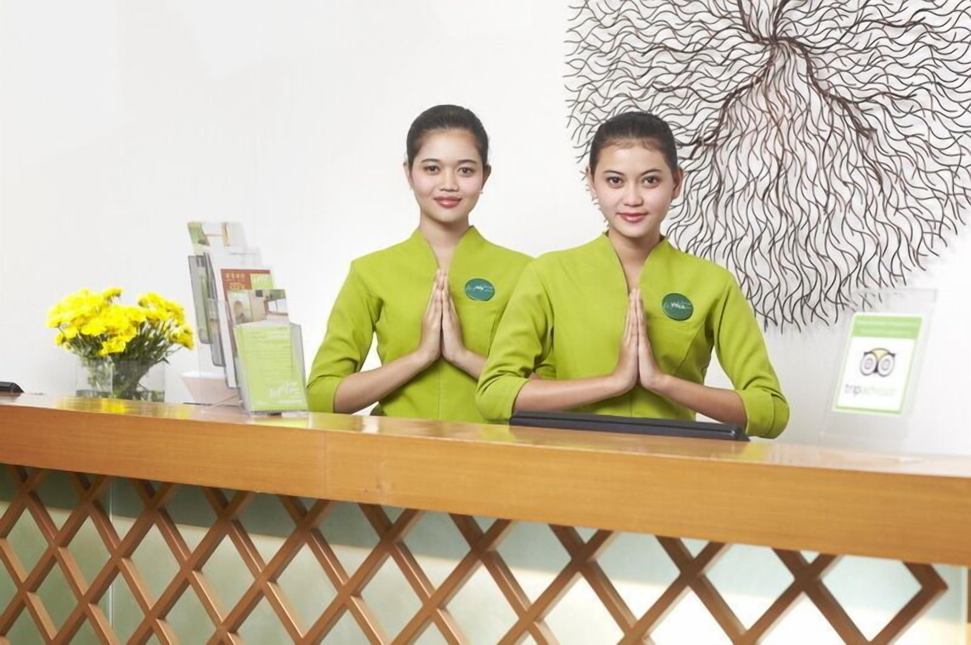 A reception desk with two staff members standing behind it, a decorative wall piece, and a vase with yellow flowers.