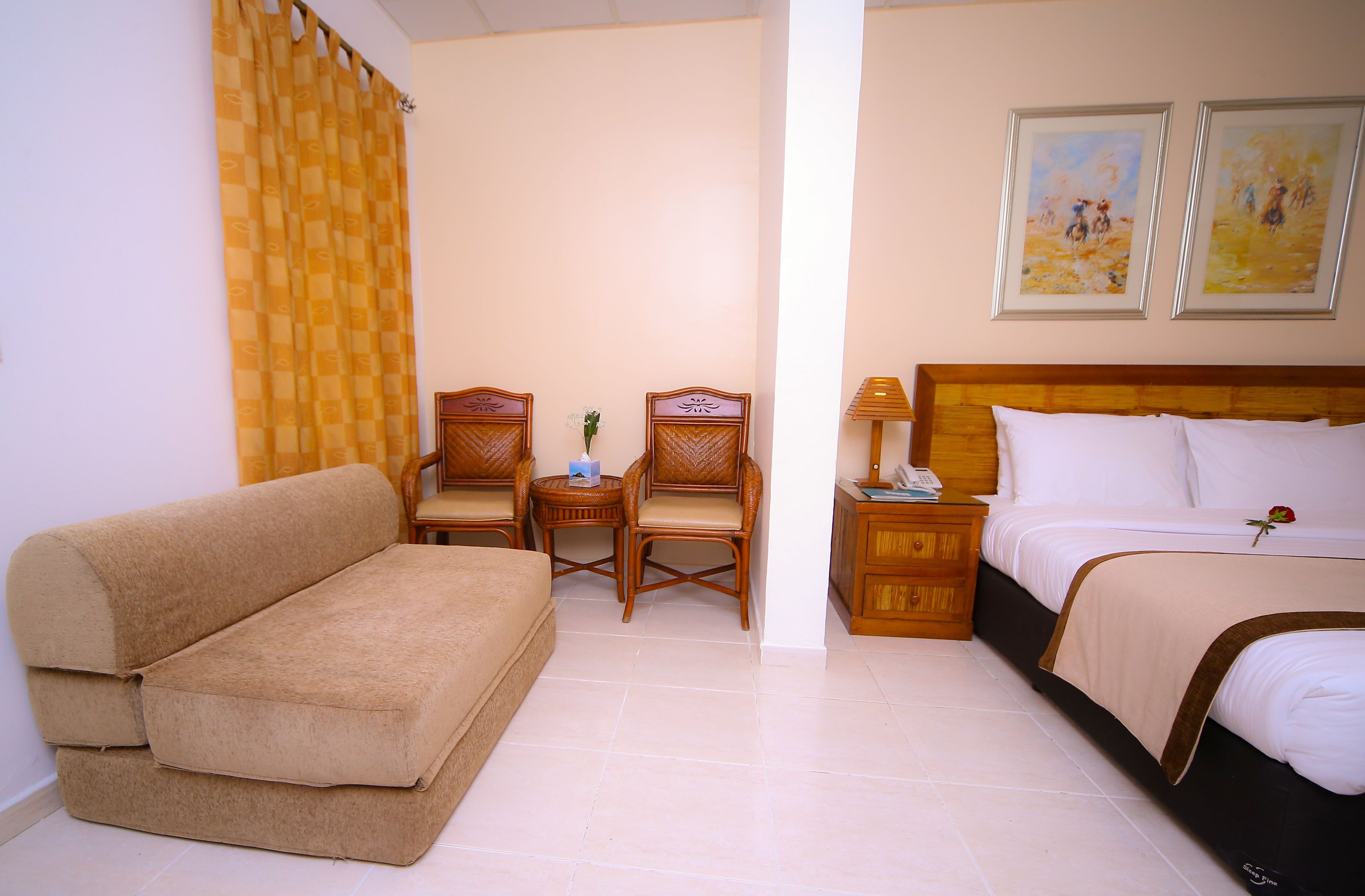Quarto de hotel com sofá, duas cadeiras, mesa de cabeceira com abajur, cama com uma rosa e duas pinturas emolduradas na parede.