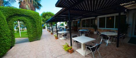 An outdoor patio area with tables and chairs, surrounded by greenery and a covered seating area.