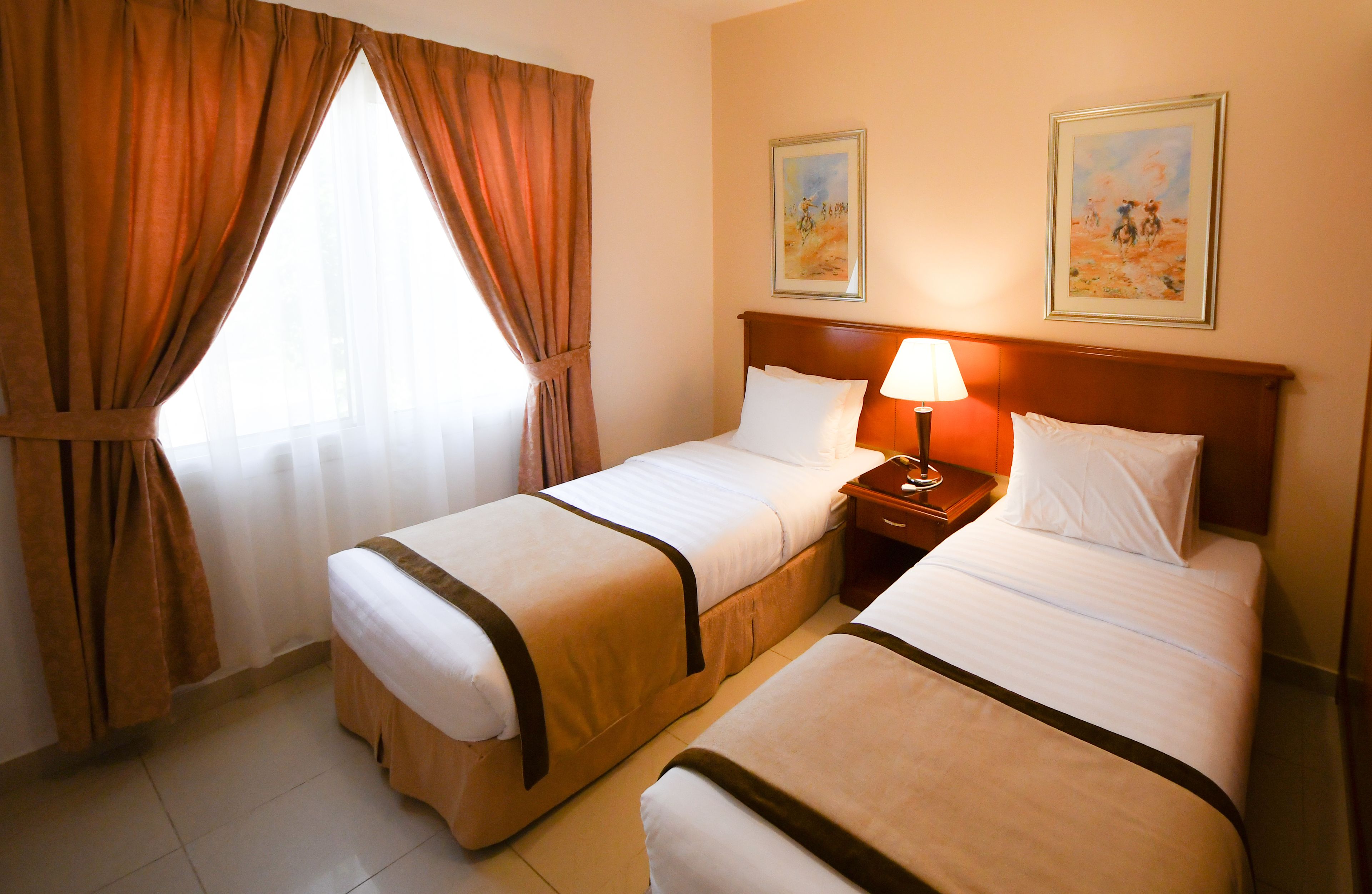 A hotel room with two beds, a nightstand with a lamp, and two paintings on the wall.