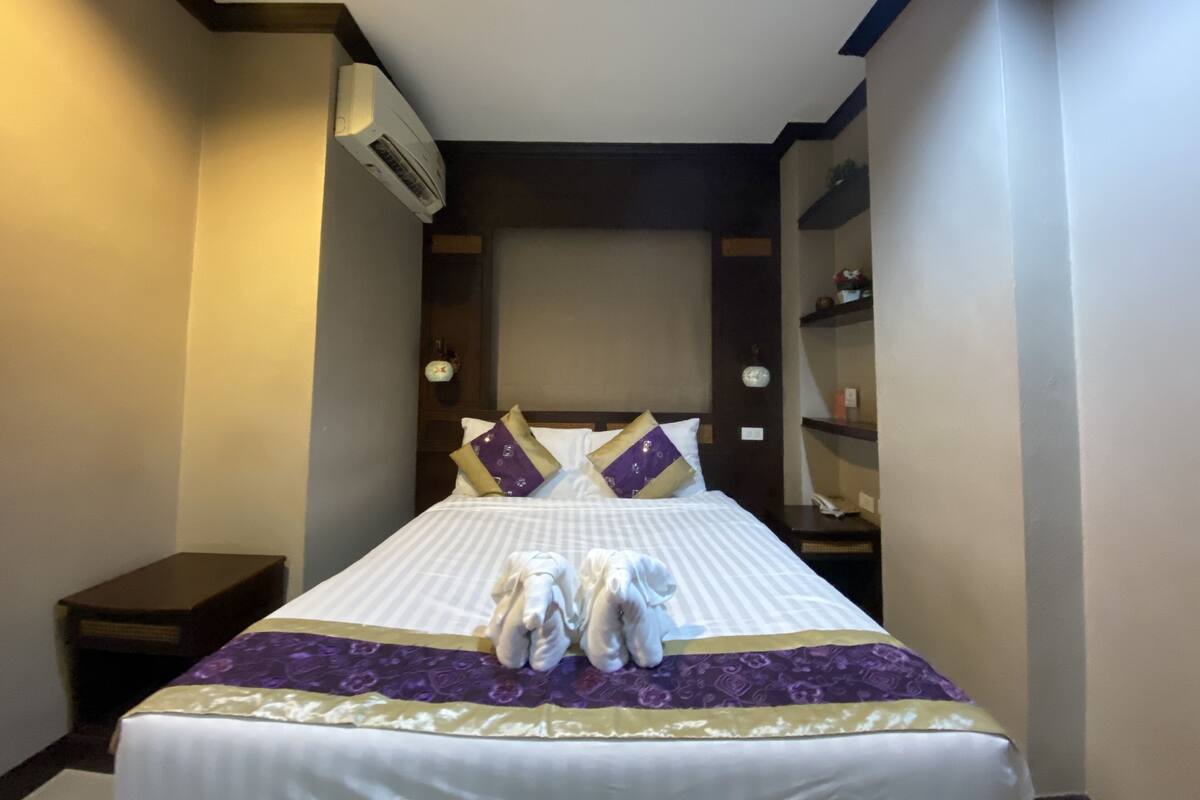 A hotel room with a neatly made bed, a bedside table, a wall-mounted air conditioner, and a small shelf with toiletries.