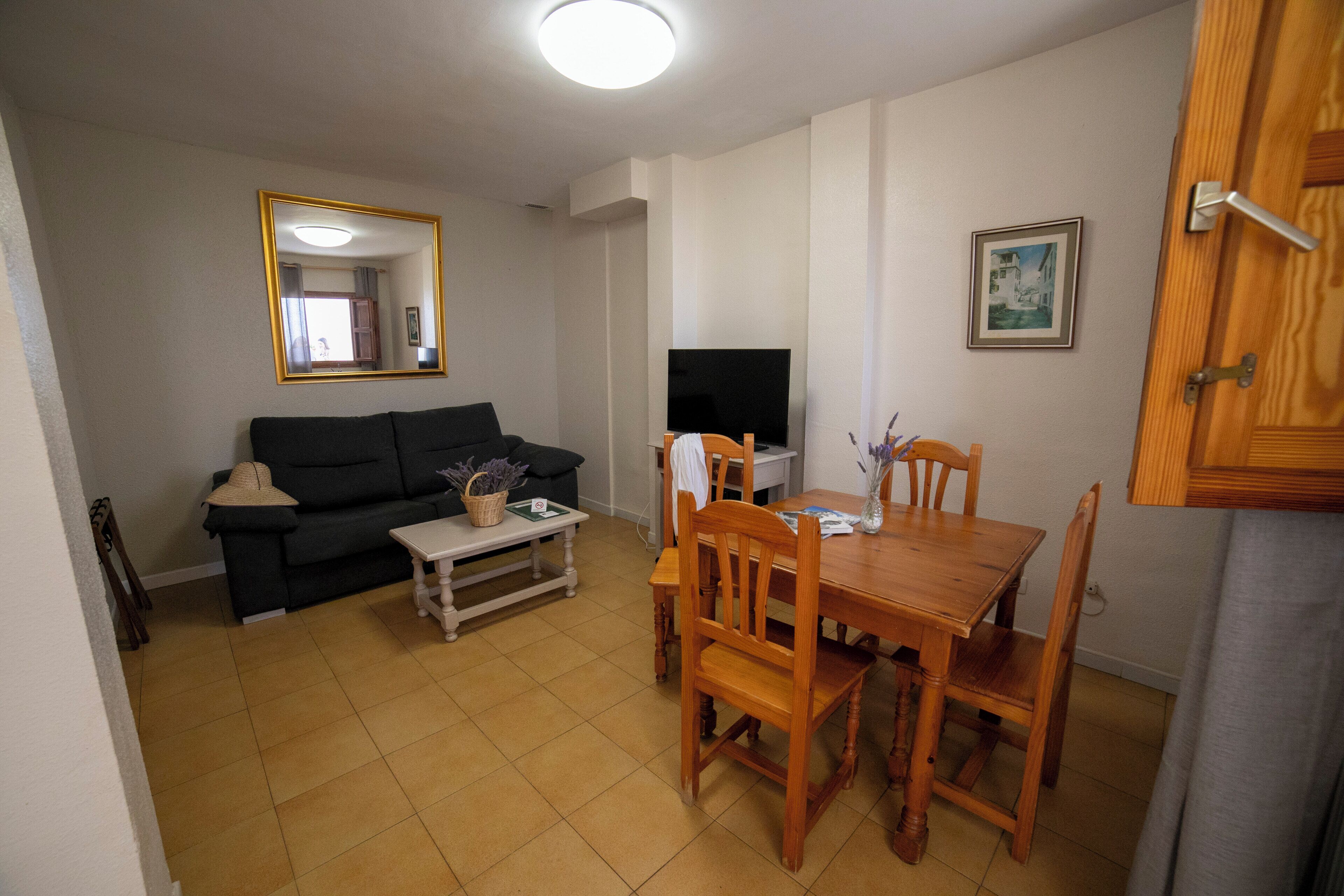 Uma sala de estar com um sofá preto, uma mesa de jantar de madeira com cadeiras, uma TV e um espelho na parede.