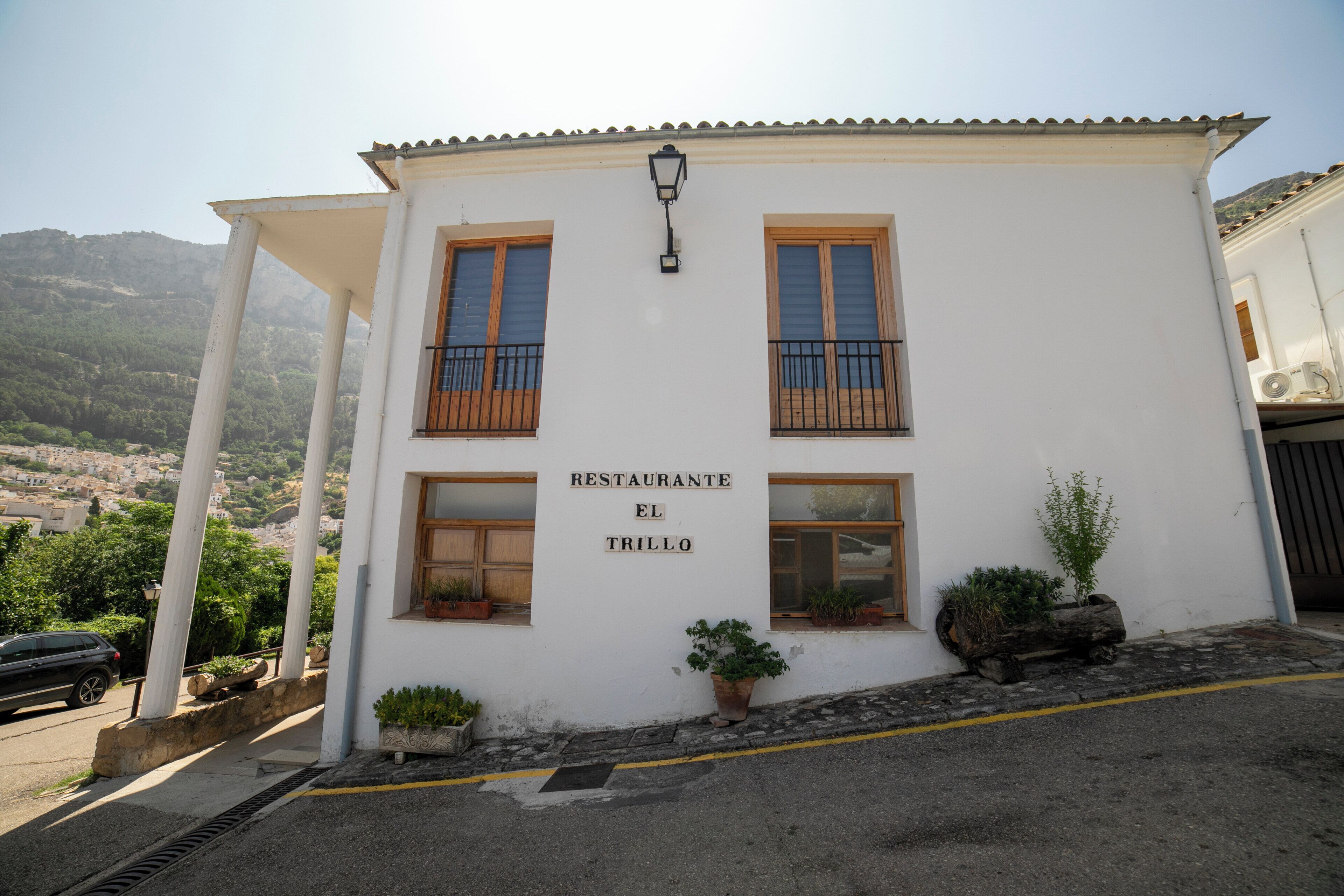 A building with a sign reading 'RESTAURANTE EL TRILLO,' windows with wooden shutters, and potted plants.