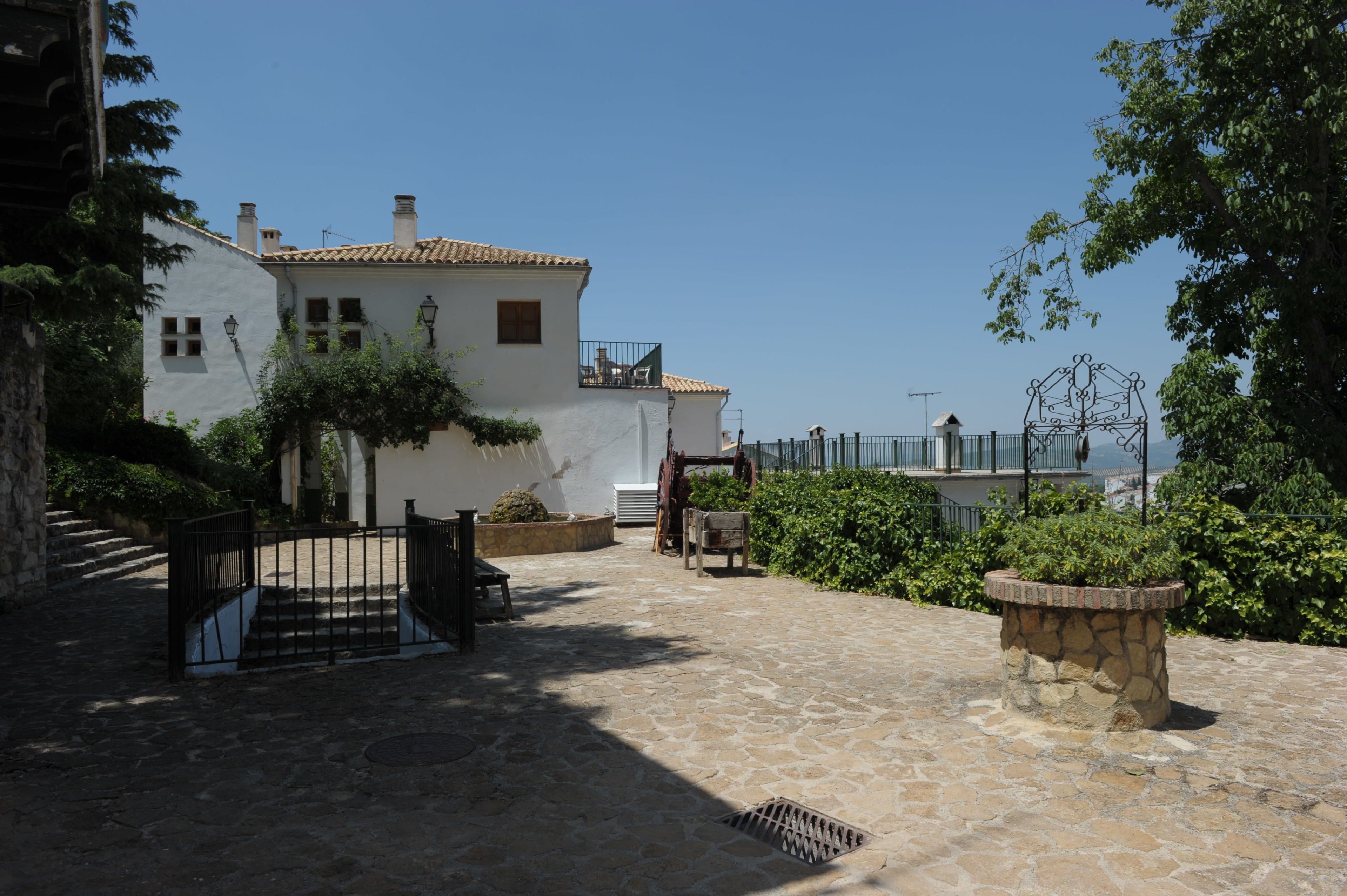 A courtyard with a stone floor, a white building with a tiled roof, and a metal gate.