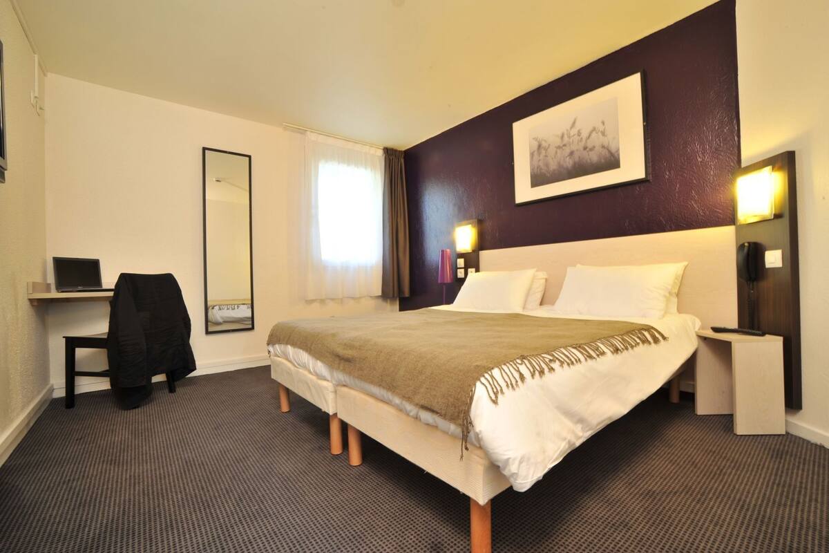 A hotel room with a bed, a desk with a laptop, a chair, a large mirror, and a painting on the wall.