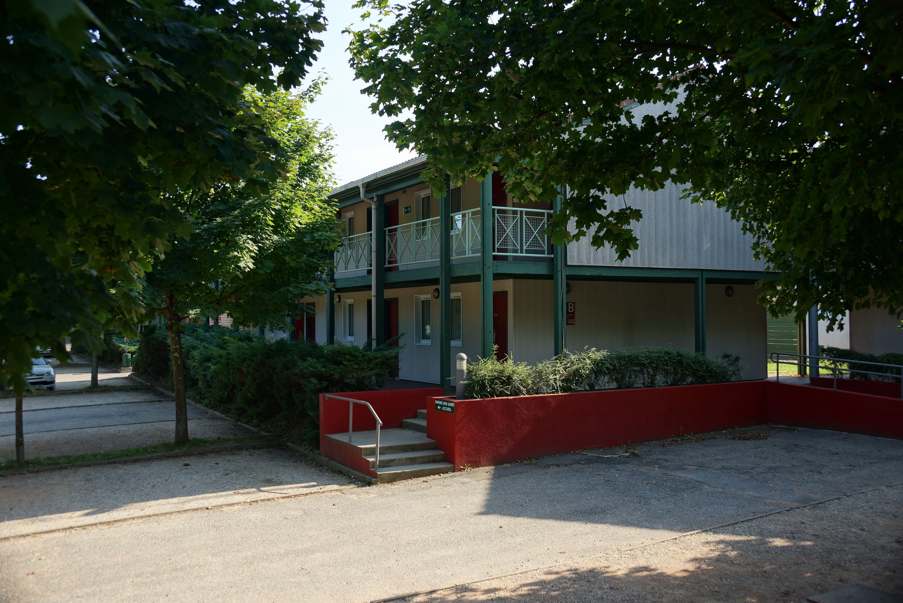 Un bâtiment à deux étages, avec une façade rouge, un balcon au deuxième étage et une allée gravillonnée.