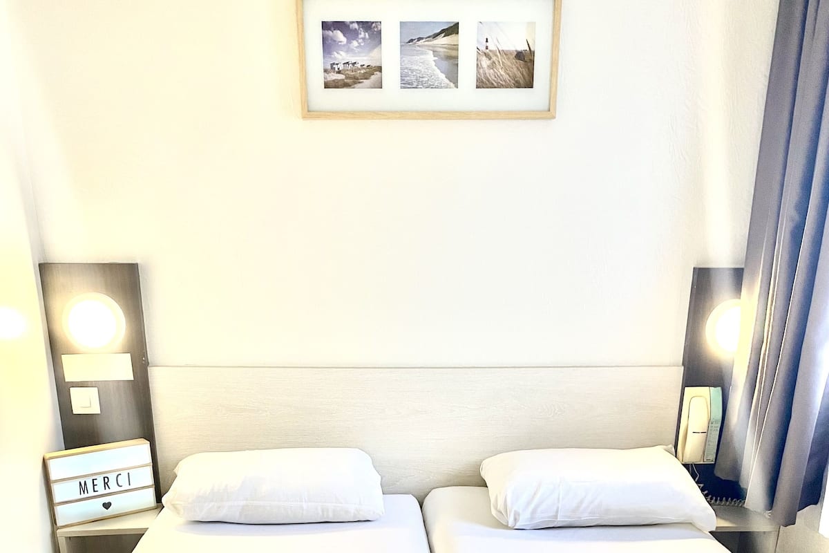 A double room with two beds, a bedside table with a lamp, a framed picture on the wall, and a sign with "MERCI" on the left side.