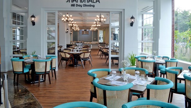 A restaurant interior with multiple dining tables and chairs, a chandelier, and a sign reading "SHAMBHALA All Day Dining".