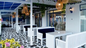 An outdoor seating area with white tables and benches, covered by a blue canopy, and hanging wicker lamps.