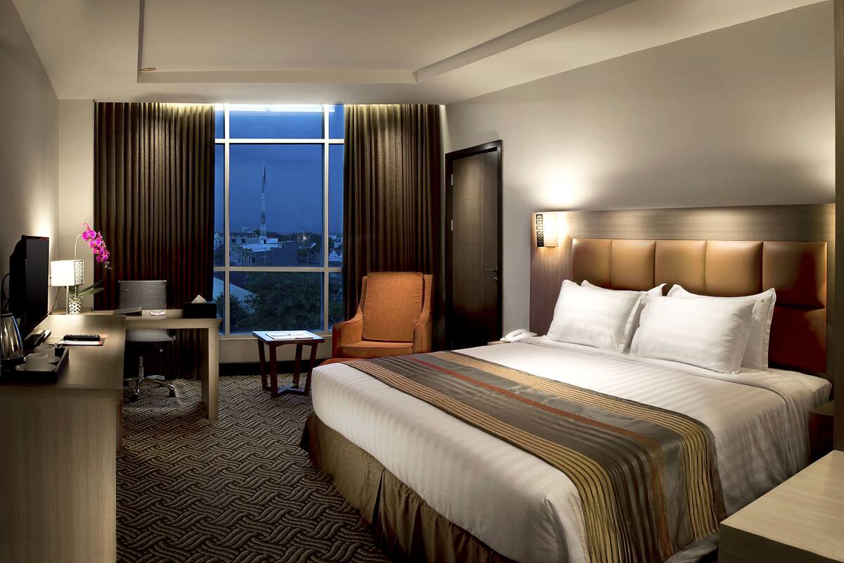 Une chambre d’hôtel avec un grand lit, un bureau avec une lampe, une chaise et une vue sur le paysage urbain par la fenêtre.