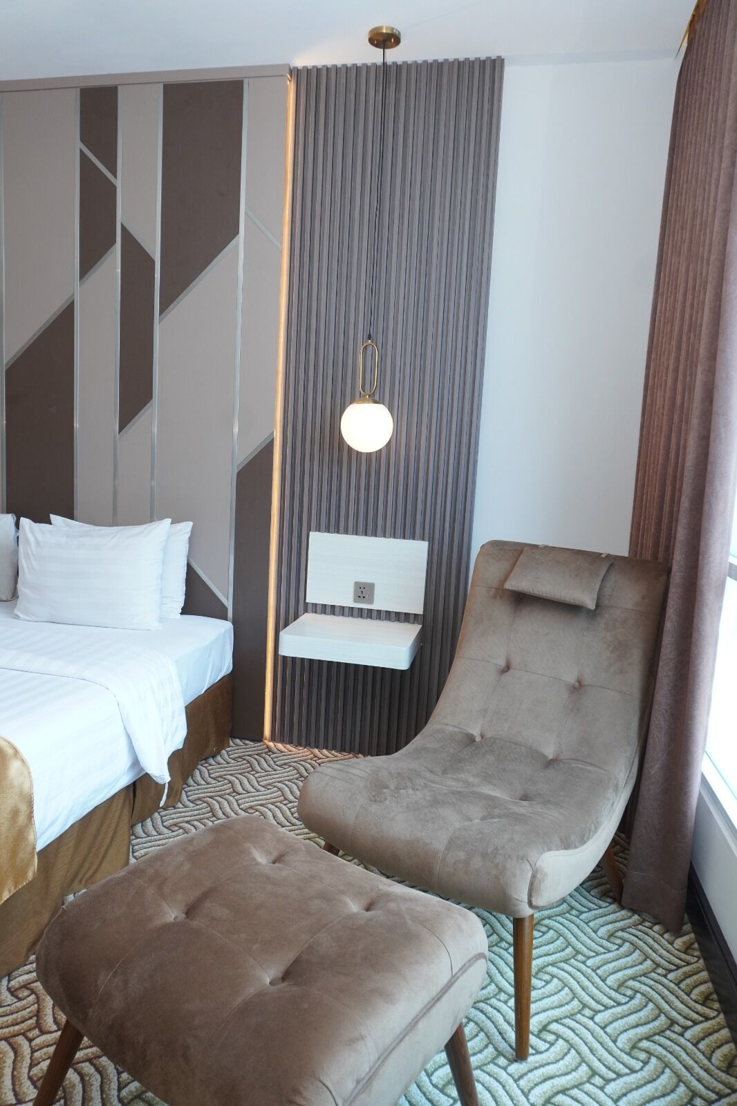 A modern hotel room with a bed, a bedside table, a chair, and a hanging light fixture.