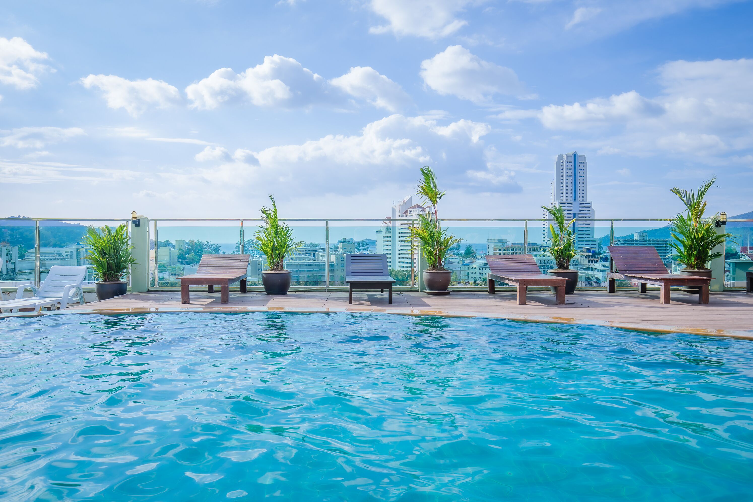 A rooftop infinity pool with lounge chairs and potted plants, overlooking a cityscape with tall buildings.