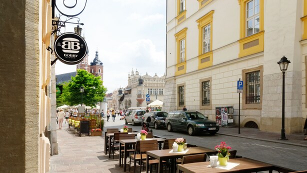 Um café ao ar livre com mesas e cadeiras, vista da rua e um edifício histórico ao fundo.