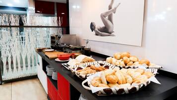 A buffet counter with various breads and pastries, a coffee machine, and a decorative wall with a black and white image.